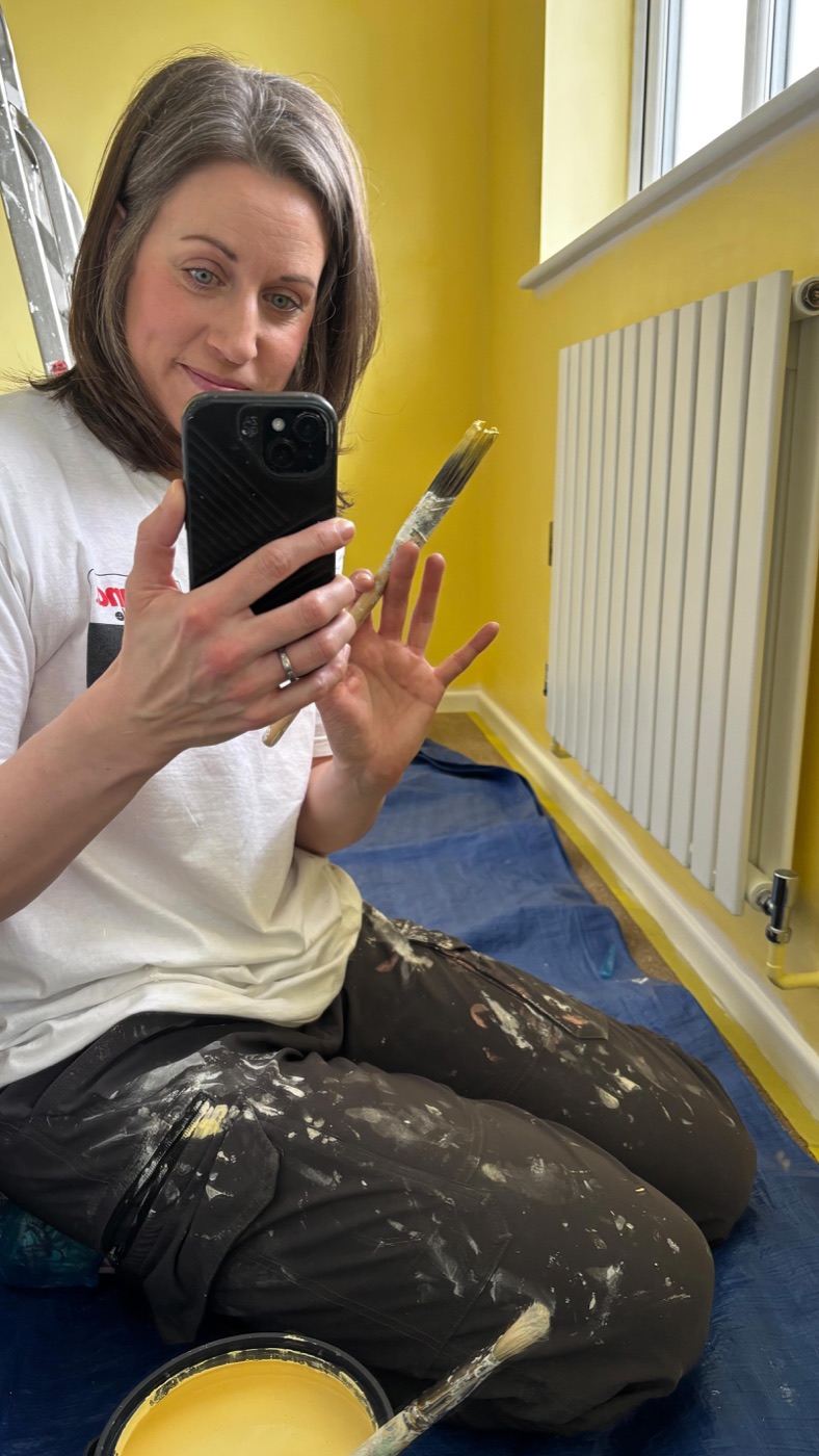 Sarah-Jane in paint-splattered workwear, smiling in a freshly painted yellow room
